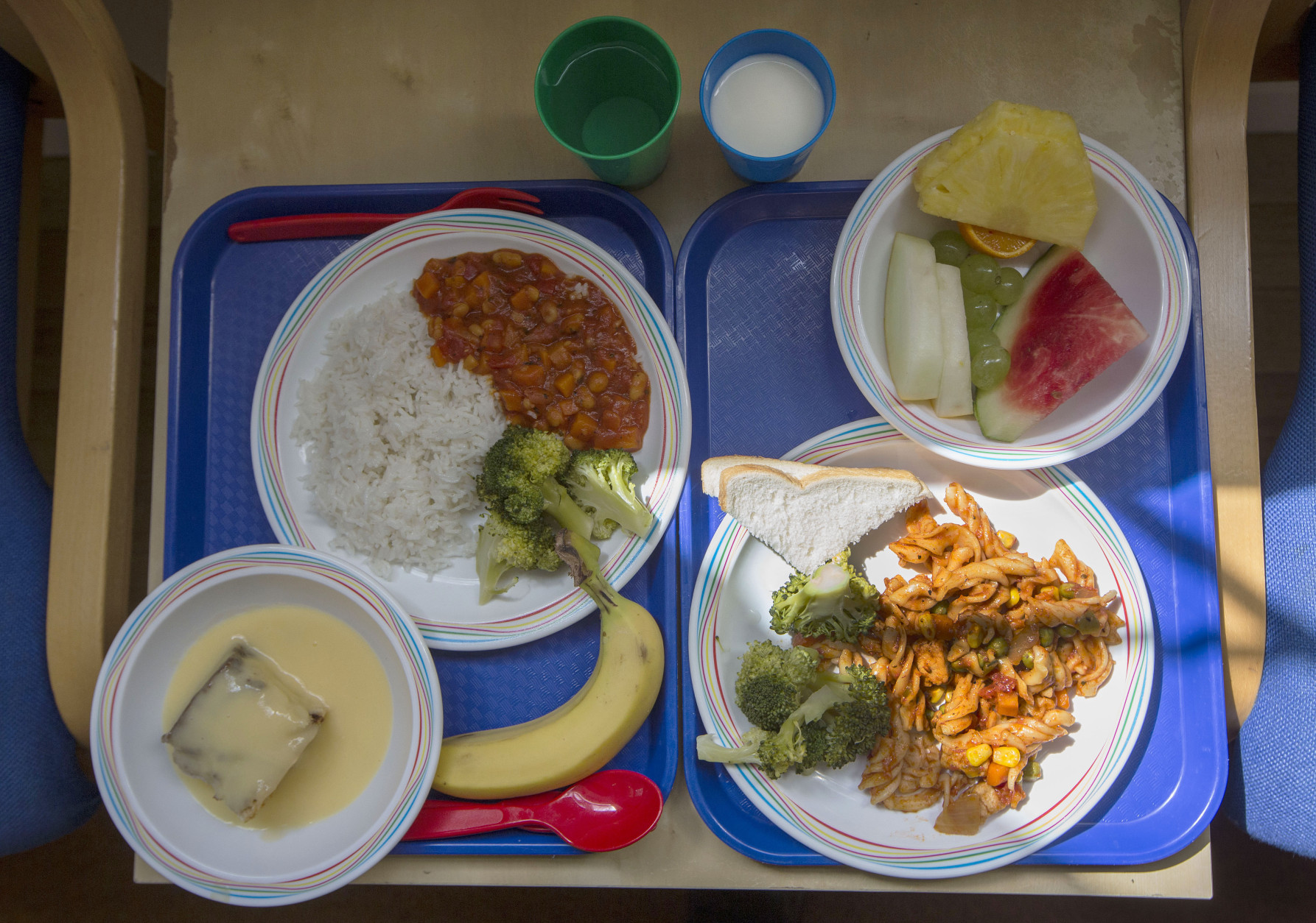 Two lunch trays at a primary school in London are served during a lunch break on Tuesday, May 6, 2014. The meal choice at right consists of pasta with fresh broccoli and slices of bread, and seasonal fresh fruit. At left are vegetable chili with rice and fresh broccoli, sponge cake with custard, and a banana. The drink options are milk and water. (AP Photo/Sang Tan)