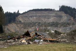 FOR USE AS DESIRED, YEAR END PHOTOS - FILE - A flag, put up by volunteers helping search the area, stands in the ruins of a home left at the end of a deadly mudslide from the now-barren hillside seen about a mile behind, Tuesday, March 25, 2014, in Oso, Wash. At least 14 people were killed in the 1-square-mile slide that hit in a rural area about 55 miles northeast of Seattle. Several people also were critically injured, and homes were destroyed. (AP Photo/Elaine Thompson, File)