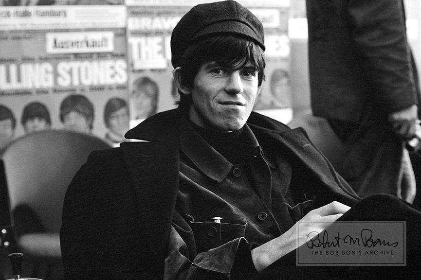 In a wonderfully meta moment, Keith Richards relaxes at a luncheon that was thrown by West German newspaper Bild Zeitung to make up for a mistake in an article that caused the Stones to threaten to cancel the rest of the tour.  The caption on a photo had misidentified Bill Wyman’s girlfriend as Chrissie Shrimpton (the younger sister of model Jean Shrimpton), who was Mick Jagger’s girlfriend which caused manager Andrew Loog Oldham to force the paper to print a retraction.  

Bob Bonis accompanied the Stones on their brief, five-city tour of West Germany - and the tour was a riot-filled affair. Former Stones bassist Bill Wyman recalls “police dogs everywhere” and crowds “estimated at between 21,000 and 23,000.” In this photo, behind Keith is a wall plastered with German Rolling Stones posters, sponsored by Bild Zeitung’s publication BRAVO. After rioting broke out at the West Berlin show on September 15, the East and West German press reprinted descriptions from Bild Zeitung of girls throwing off their underwear in ecstasy - in an effort to censor American and British influences, just as they had done with Elvis Presley in 1956.