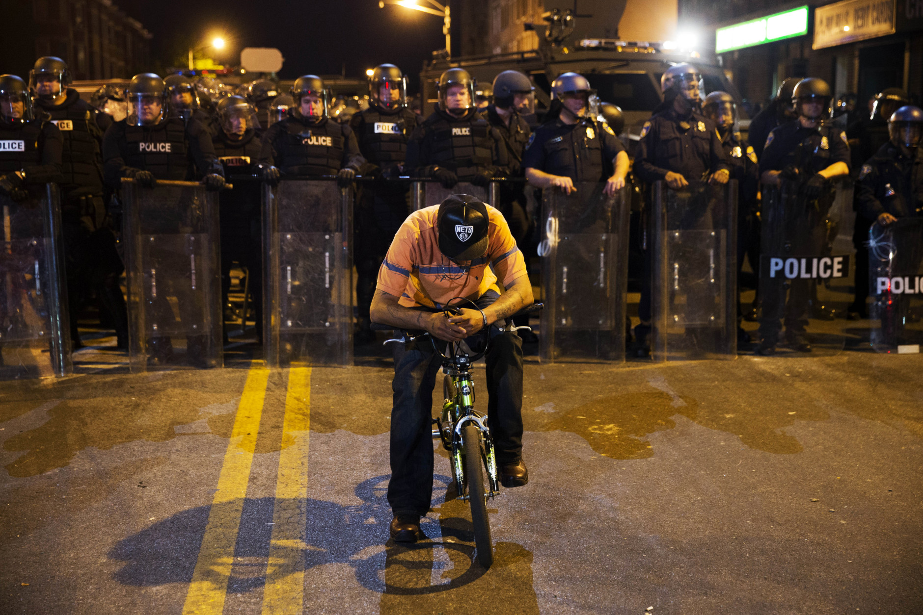 A man sits on a bicycle in front of a line of police officers in riot gear as part of a community effort to disperse the crowd ahead of a 10 p.m. curfew in the wake of Monday's riots following the funeral for Freddie Gray, Tuesday, April 28, 2015, in Baltimore. (AP Photo/David Goldman)