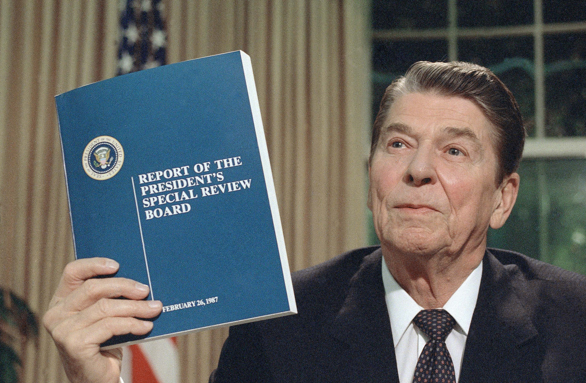 President Ronald Reagan holds up a copy of the Tower Commission report on the Iran-Contra affair, while posing for photographers after his nationally televised speech from the Oval Office in Washington, Aug. 13, 1987. Reagan said he was mad as a hornet about damage to his administration from the Iran-Contra affair. (AP Photo/Ron Edmonds)