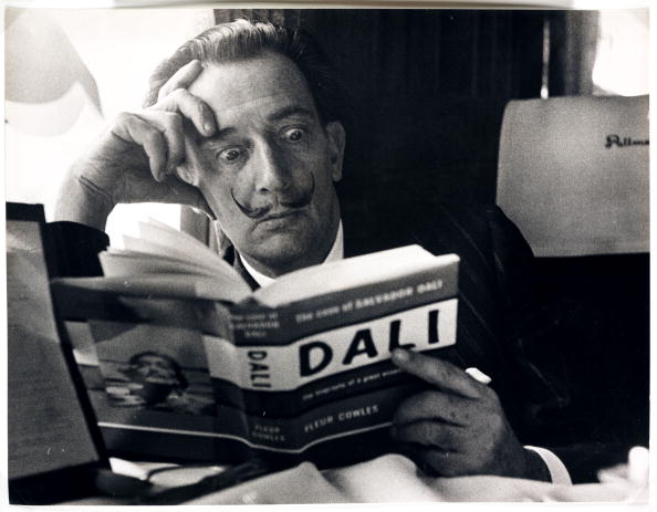 UNITED KINGDOM - NOVEMBER 21:  Salvador Dali reading his biography, 6 May, 1959. A photograph of the Spanish artist Salvador Dali (1904-1989), taken by Terry Fincher for the Daily Herald newspaper. Dali is reading Fleur Cowles' book 'The Case of Salvador Dali' (1959), whilst on a train from Folkestone, having travelled from France. Cowles' book was an authorised biography of Dali. With a studied expression of shock on his face, Dali enjoys the photo opportunity. One of the most famous, charismatic and notorious artists of the twentieth century, Dali devoted himself to drawing and painting from an early age. Hugely influential as a Surrealist, one of his most famous paintings is 'The Persistence of Memory'. By the 1950s Dali had turned to demonstrating scientific, historical and religious themes in his work. In later life he founded both the Dali Theatre-Museum, Figueres, Spain and the Gala-Salvador Dali Foundation, Pubol Castle, Spain - the latter to manage his legacy.  (Photo by Daily Herald Archive/SSPL/Getty Images)