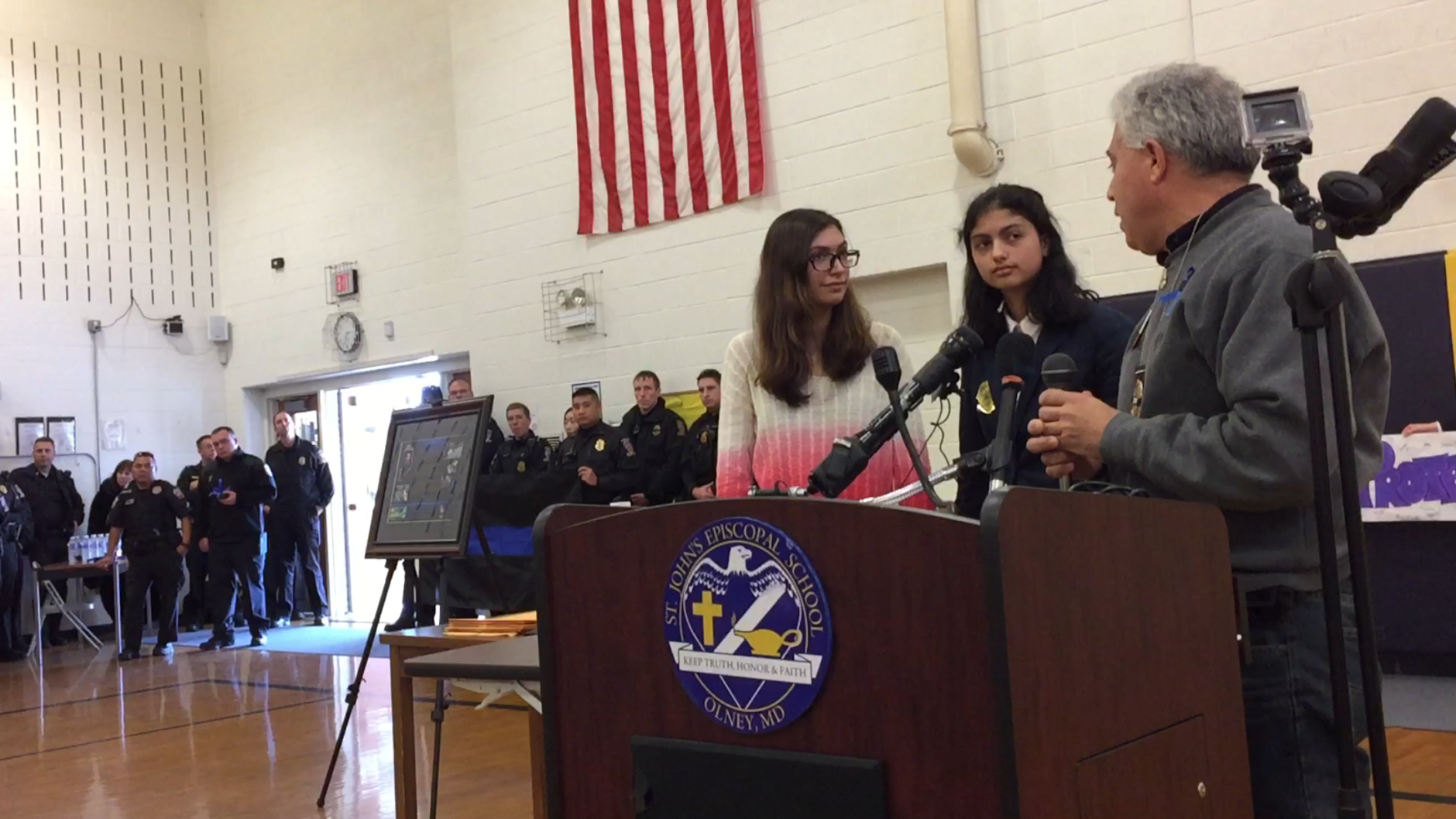 Standing with her sister Katherine-Aria Close, seventh grader Alexandra Close, center, is thanked by Rick Leotta for her poem commemorating the day she and classmates honored Leott'a son, fallen officer Noah Leotta along his funeral procession. Officers in background also attended the ceremony in which Close and her school were given a plaque of appreciation by Leotta and the police department.(WTOP/Kristi King)