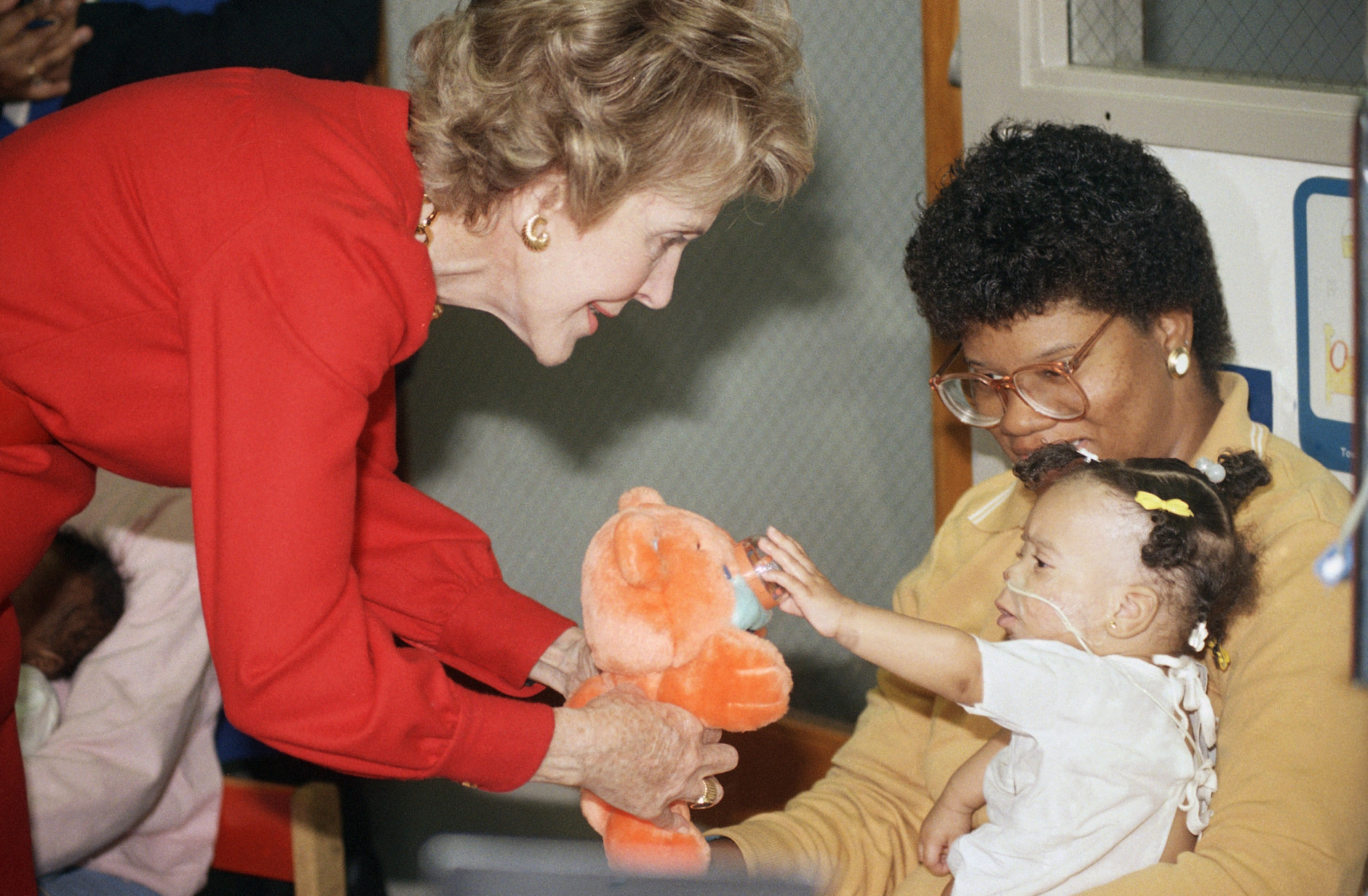 Eleven month old Nicole Seay reaches out to touch the nose of a teddy bear held by first lady Nancy Reagan on Wednesday, Dec. 14, 1988 at the Children's Hospital in Washington.   Nicole is held by her mother Corinthia.    Mrs. Reagan makes an annual holiday visit to the hospital to greet the children. (AP Photo/Rick Bowmer)
