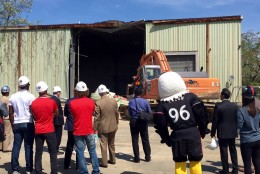 District and team leaders, and the DC United mascot, look on as a backhoe tears down the empty structure on the stadium site. (WTOP/Megan Cloherty)
