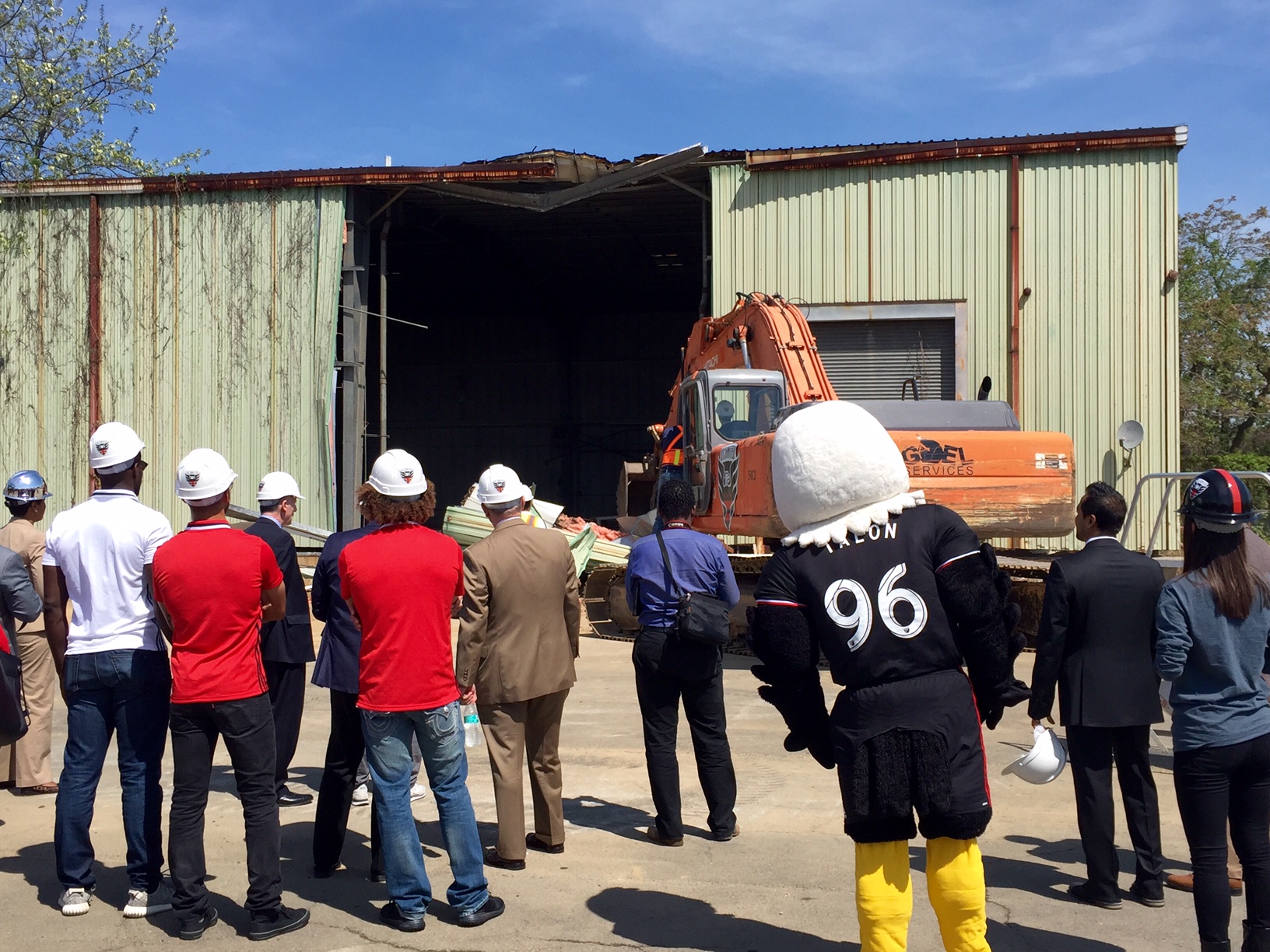 District and team leaders, and the DC United mascot, look on as a backhoe tears down the empty structure on the stadium site. (WTOP/Megan Cloherty)