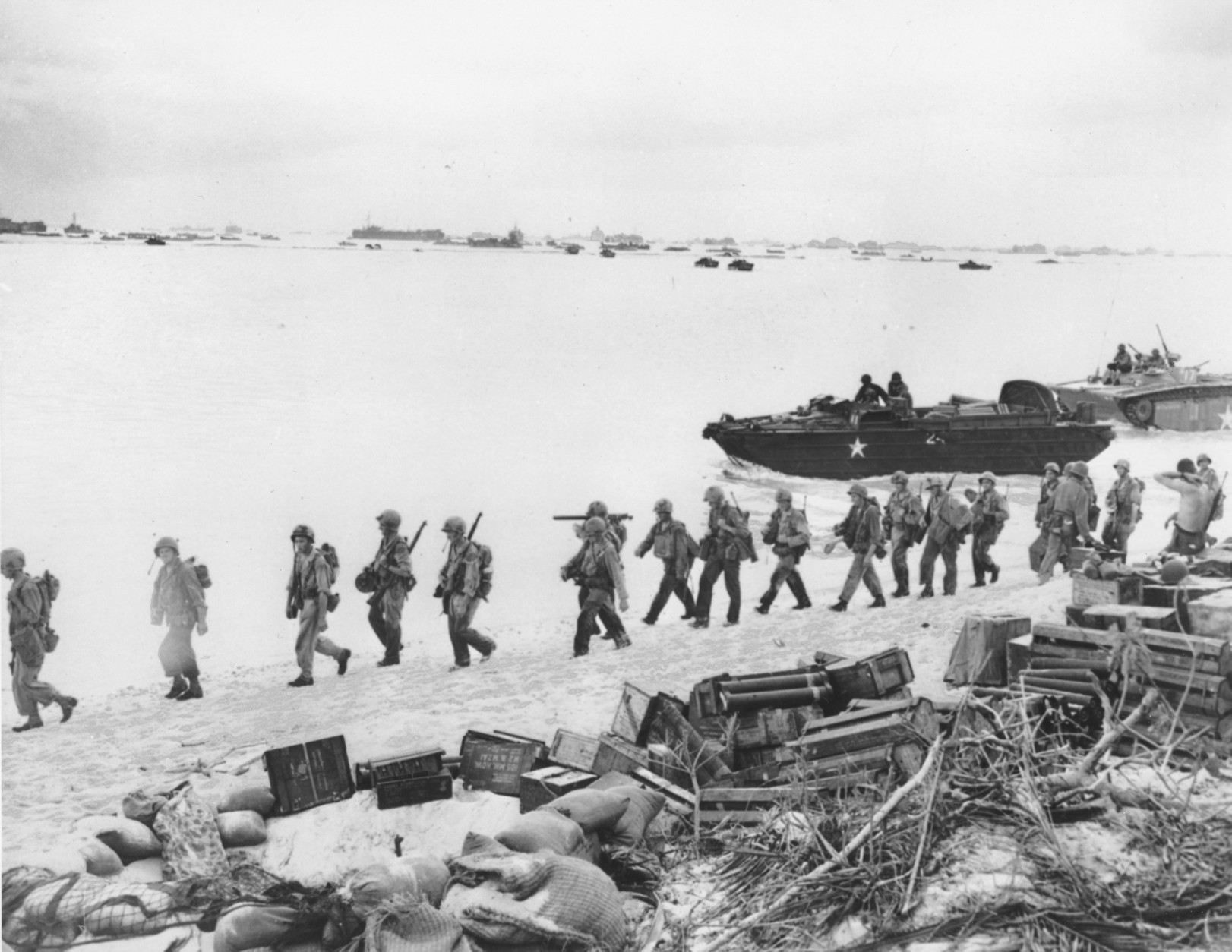 American troops march along a beach on Saipan in the Mariana Islands after debarking from amphibious tanks during invasion operations against Japanese forces in June 1944 during World War II.  Army supplies are brought ashore, foreground.  On the horizon are various ships that make up the invasion force.  (AP Photo)