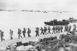 American troops march along a beach on Saipan in the Mariana Islands after debarking from amphibious tanks during invasion operations against Japanese forces in June 1944 during World War II.  Army supplies are brought ashore, foreground.  On the horizon are various ships that make up the invasion force.  (AP Photo)