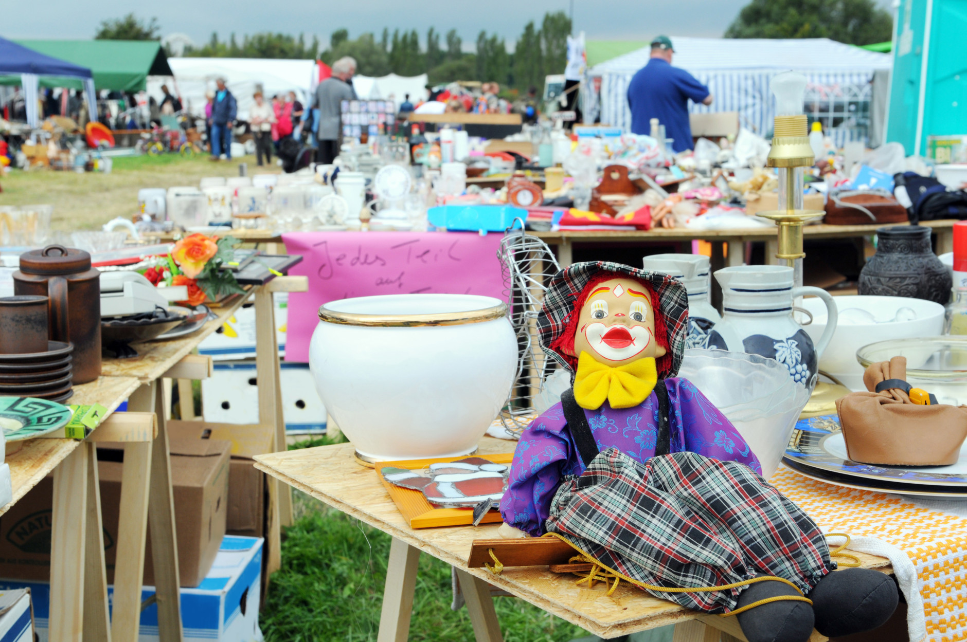 It's a typical impression of Flea Market at Havelberg (Saxony-Anhalt, Germany), that takes place every year around the first September Weekend. In front tables with colorful clown marionette for puppet show and dishware. in background group of visitors taking a look on products. dark clouds on shy. in background German words "jedes Teil nur"... its English every item just...
