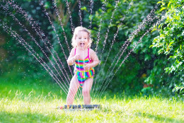 Funny laughing little girl in a colorful swimming suit running though garden sprinkler playing with water splashes having fun in the backyard on a sunny hot summer vacation day