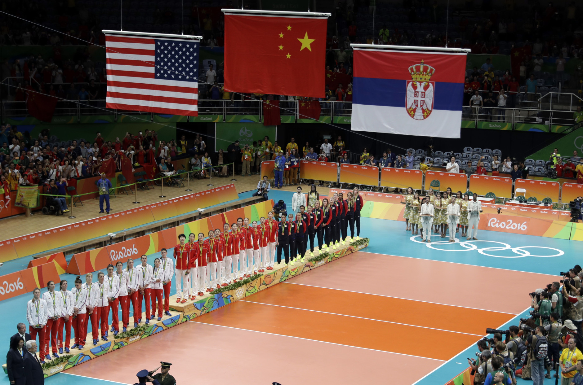 The teams of China, center, Serbia, left, and the United States watch as their flags are raised during the awards ceremony for women's volleyball at the 2016 Summer Olympics in Rio de Janeiro, Brazil, Saturday, Aug. 20, 2016. China was awarded gold while Serbia was awarded silver and the United States took bronze. (AP Photo/Jeff Roberson)