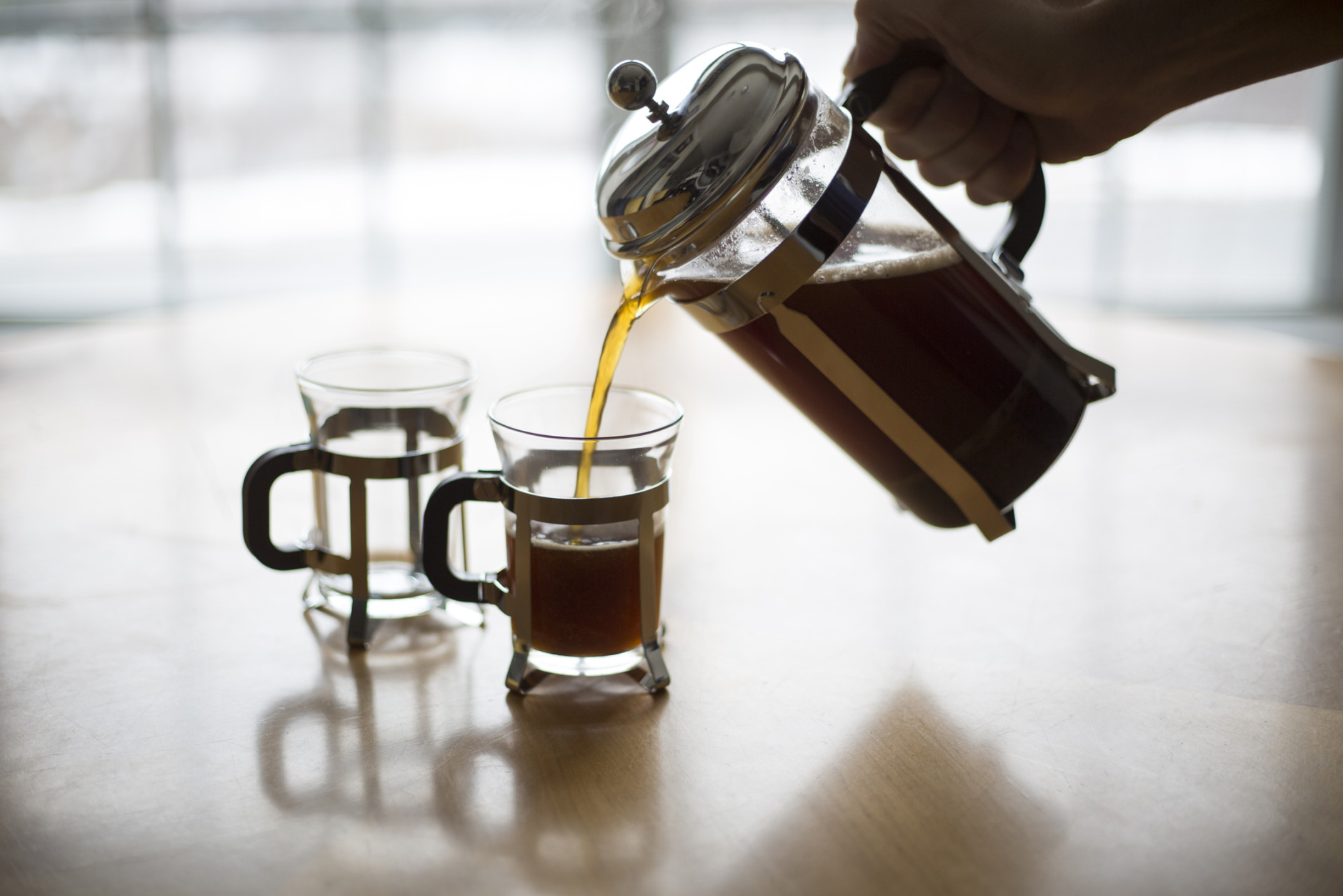 A back lit image of coffee being poured into small coffee mugs from a French Press with the light source being a window behind the table.