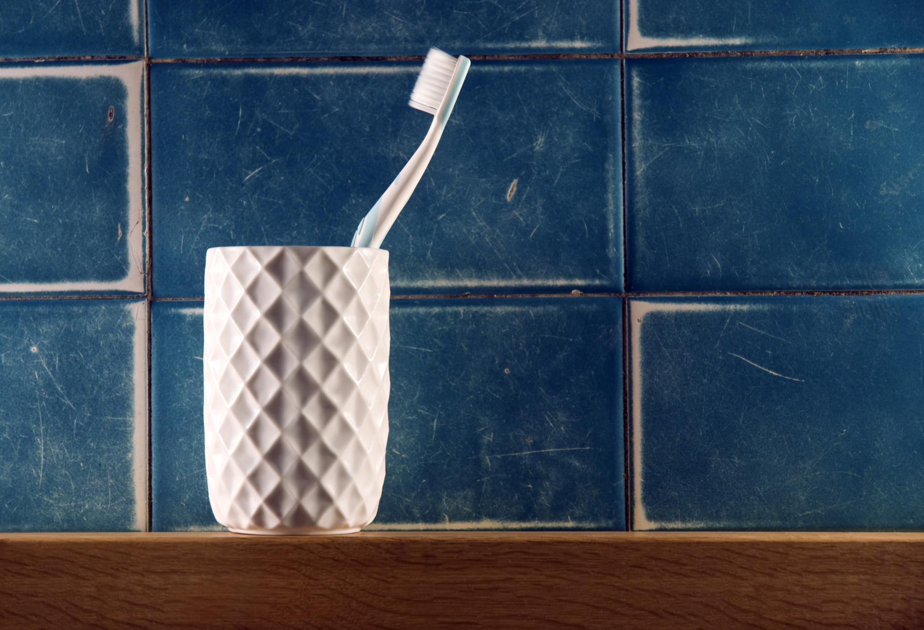Toothbrush in a container sitting on a wooden shelf in front of a tiled wall.