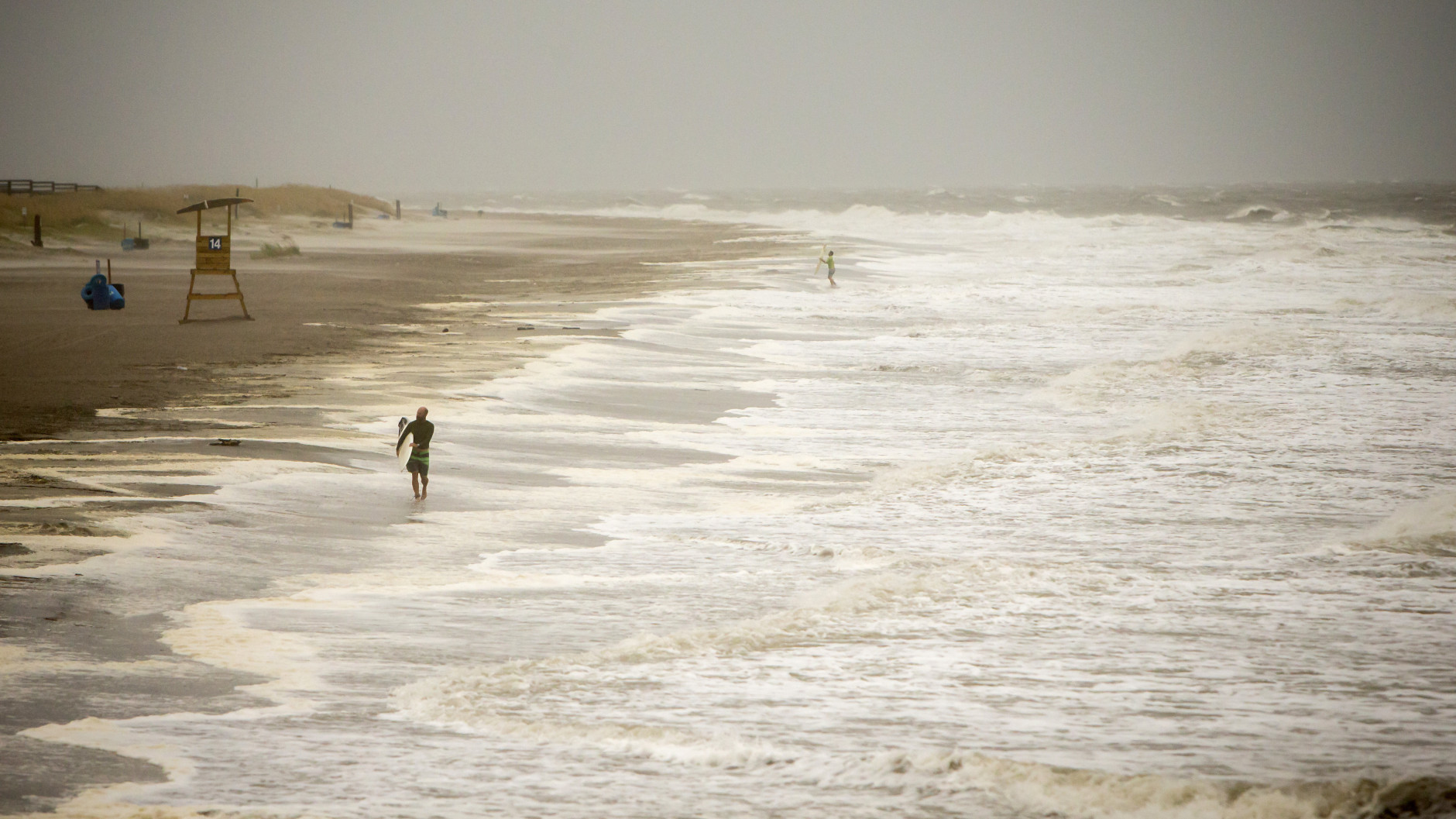 A surfer walks the beach while looking for waves from the surge of Hurricane Hermine, Friday, Sept. 2, 2016, off the coast of Tybee Island, Ga.  Hermine was downgraded to a tropical storm after it made landfall, as it moves over Georgia, but the U.S. National Hurricane Center says winds are increasing along the Southeast coast and flooding rains continue.  (AP Photo/Stephen B. Morton)