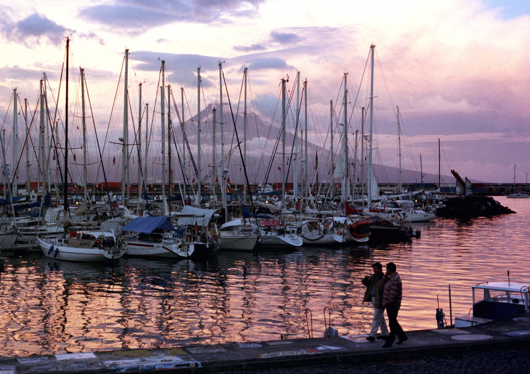 Two yachtsmen walk along the Horta harbor at sunset on the Azores island of Faial, June 18, 1999. The 7,755-foot-high (2,350 meter) volcanic peak of Pico island rises in the background. Transatlantic yachtsmen are increasingly being joined by tourists to the nine small islands of the Azores, seeking a natural, unspoiled escape from the stress of city life. (AP Photo/Armando Franca)