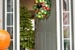 Open doorway to an inviting Christmas scene with a colorful decorated wreath hanging on the wall and pumpkins on the porch in the foreground