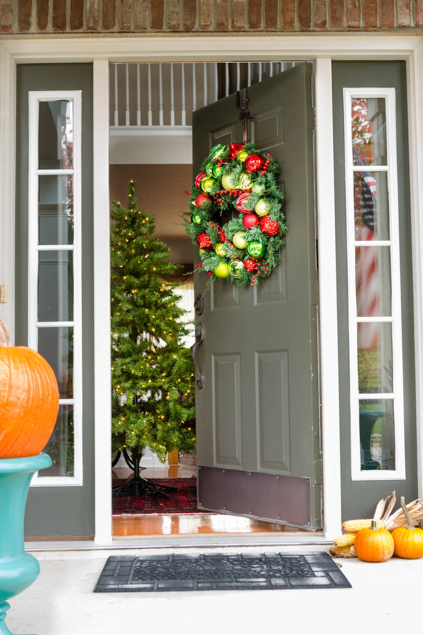 Open doorway to an inviting Christmas scene with a colorful decorated wreath hanging on the wall and pumpkins on the porch in the foreground