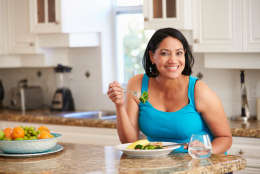 Overweight Woman Eating Healthy Meal in Kitchen Whilst Looking At Camera Smiling
