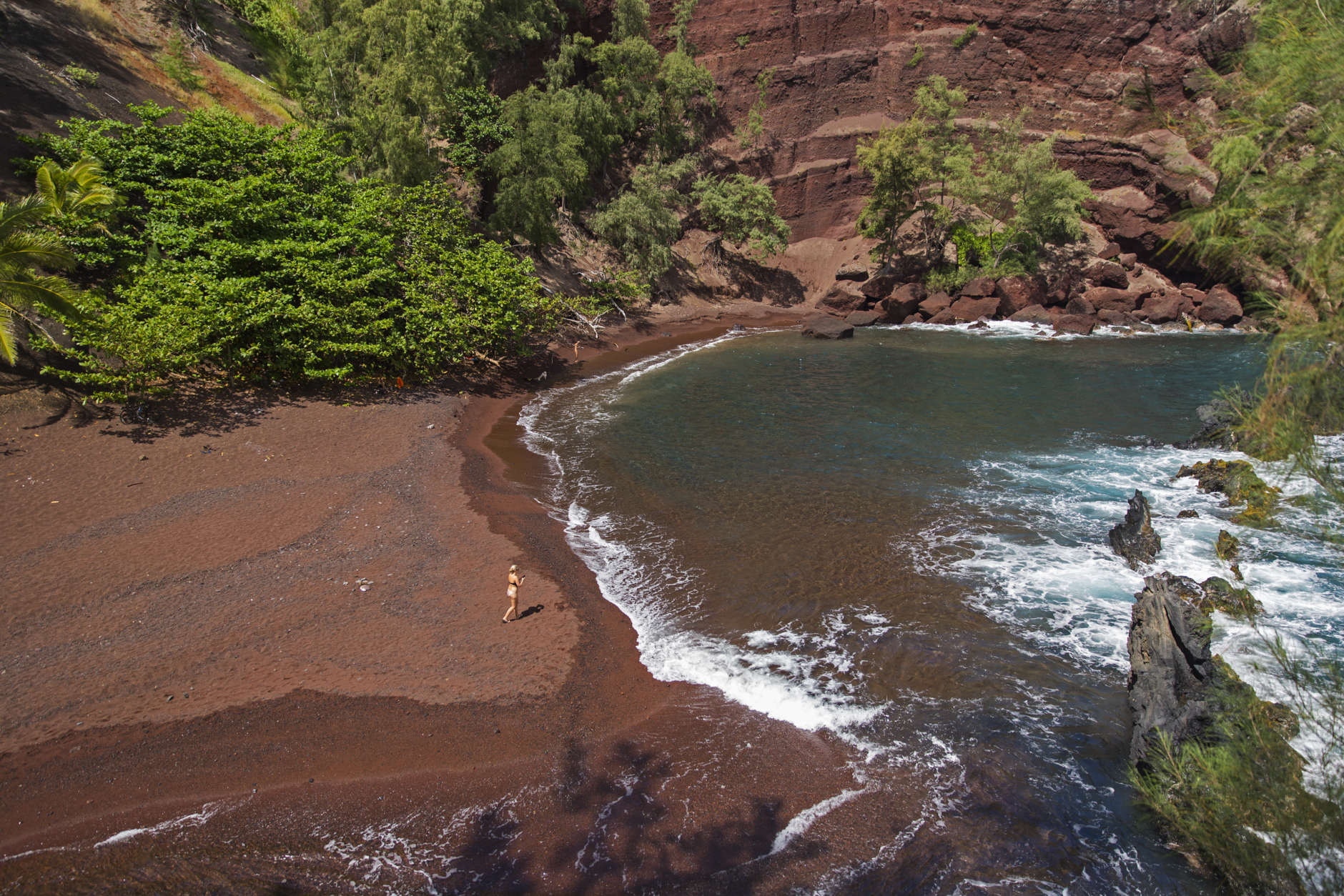 FILE - In this Sept. 24, 2014 file photo, a woman walks on the red sand beach at Kaihalulu Bay in Hana, Hawaii. Towering red cinder cliffs surround the bay, and the blue ocean swirls along the red sandy shore. A large lava rock reef juts out in the bay slightly protecting the beach from harsh waves, but swimming is not advised. (AP Photo/Marco Garcia, File)