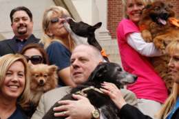 Maryland Gov. Larry Hogan, a dog-lover, holds Mabel, a rescue dog, before signing bills into law. Among the bills he signed, one that helps animal welfare agencies recover the costs associated when they rescue animals from hoarding cases.  That's Oreo behind him, giving out smooches. Over his shoulder is fluffy, little Gem, and off to the right with a big grin, is Rocky. (WTOP/Kate Ryan)