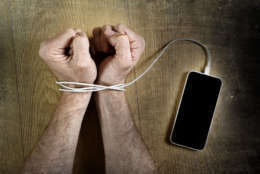 man hands trapped and wrapped on wrists with mobile phone cable as handcuffs in smart phone networking and communication technology addiction concept