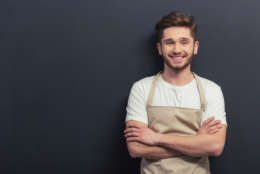 Handsome young man in apron is looking at camera and smiling, standing with crossed arms against blackboard