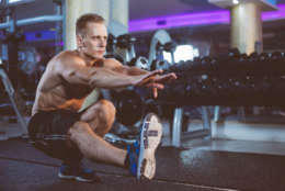 Attractive young blond fitness man doing one leg squat exercise in gym. Background of gym equipment.