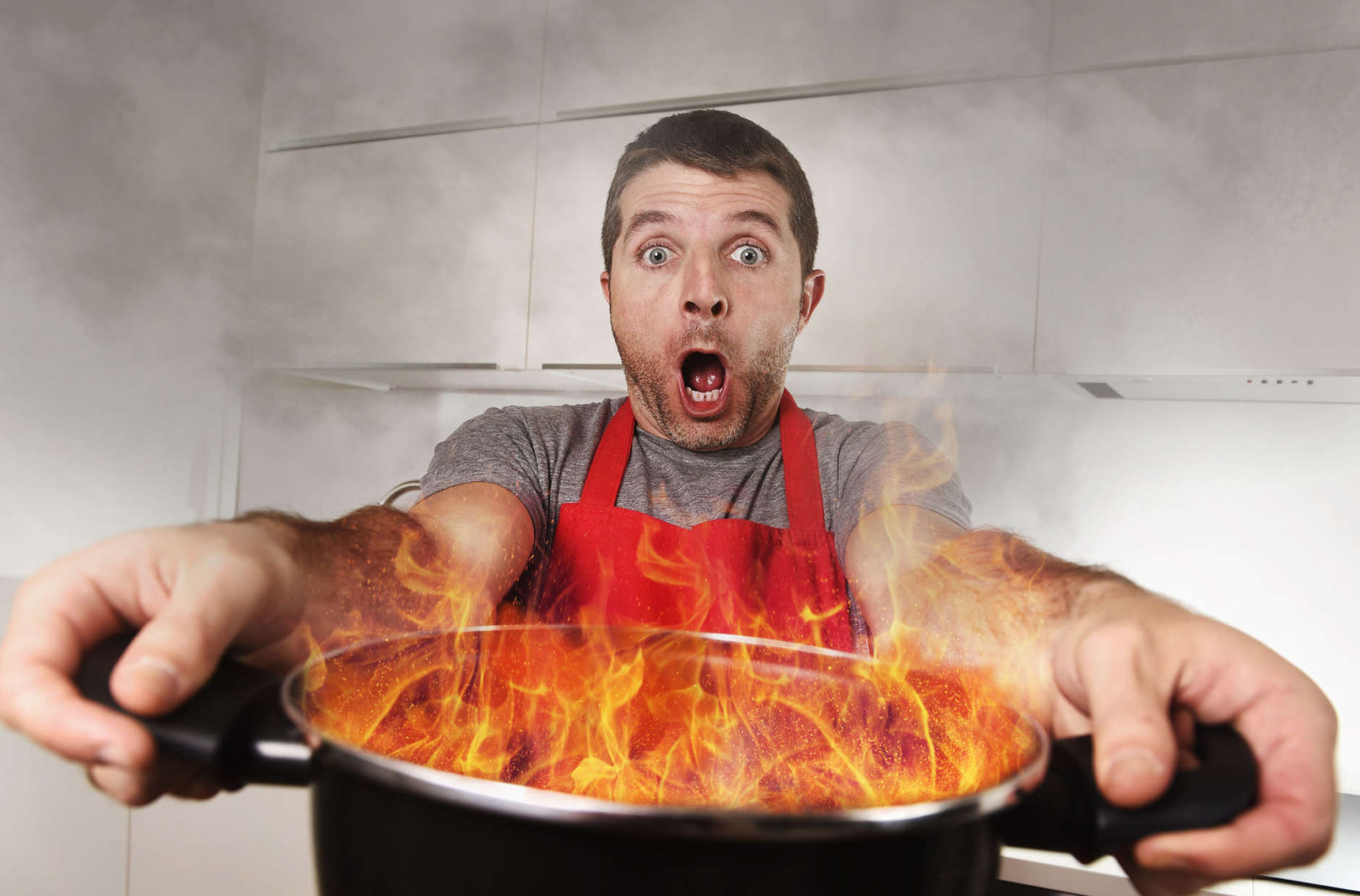 young inexperienced home cook with apron holding pot burning in flames with stress and panic face expression in fire in the kitchen and cooking wrong concept