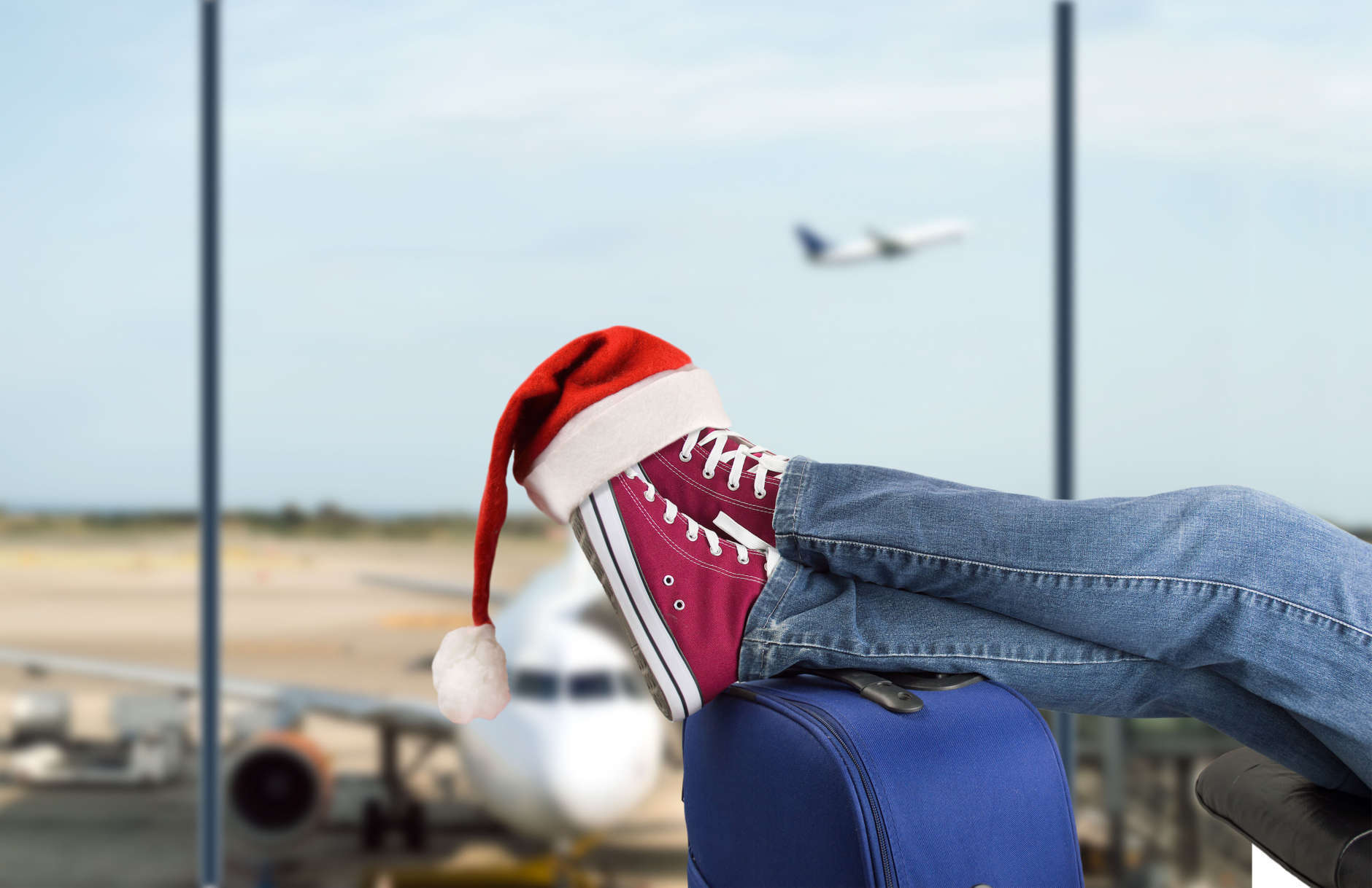 young boy with his legs over the suitcase waiting at the airport with santa hat on the feet
