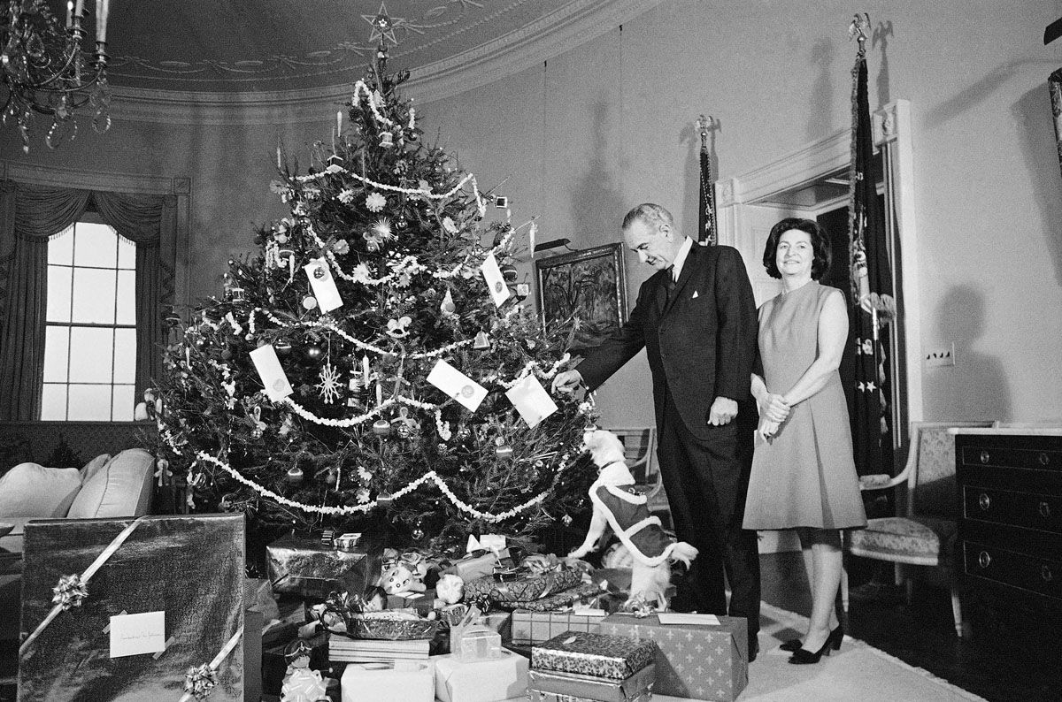 FILE - In this Dec. 24, 1967, file photo President Lyndon Johnson,  Lady Bird Johnson, and Yuki, the White House pet, pose beside the family Christmas tree, a Norway spruce, in the Yellow Oval Room on the second floor of the White House in Washington. After President John F. Kennedy's assassination on Nov. 22, 1963, a month of mourning was declared. But on the evening of Dec. 22, Johnson lit the National Christmas Tree behind the White House, and the next morning the black mourning crepe that had been draped over White House doorways and chandeliers was replaced with holly, wreaths and mistletoe. Lady Bird Johnson later wrote, "I walked the well-lit hall for the first time with the sense that life was going to go on, that we as a country were going to begin again."  (AP Photo/File)