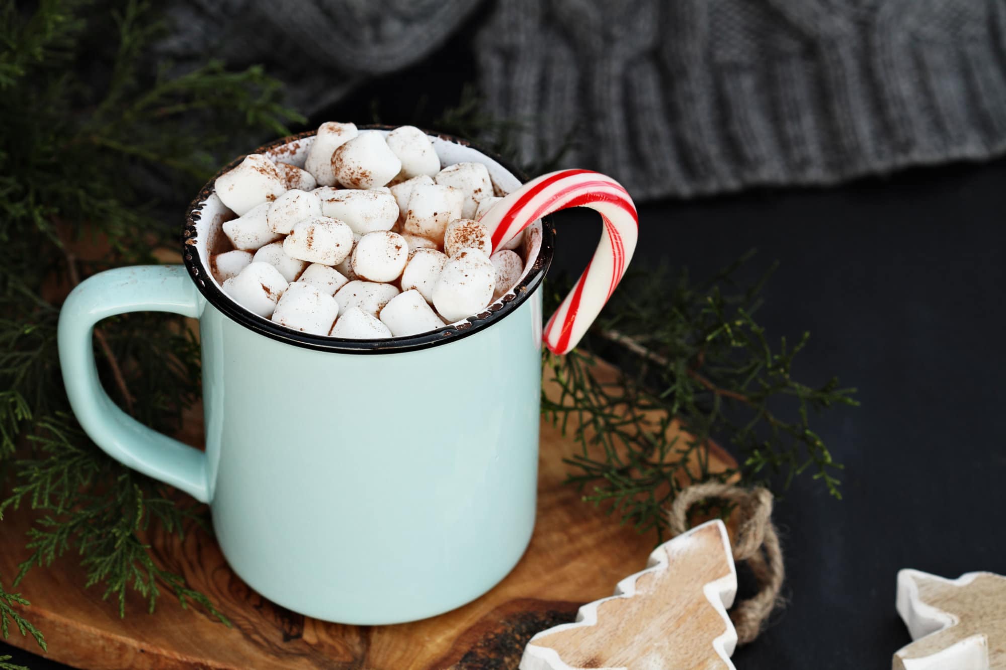 Enamel cup of hot cocoa drink with marshmallows and candy cane against a rustic background with beautiful wood Christmas tree ornaments and a grey scarf. Perfect winter time treat.