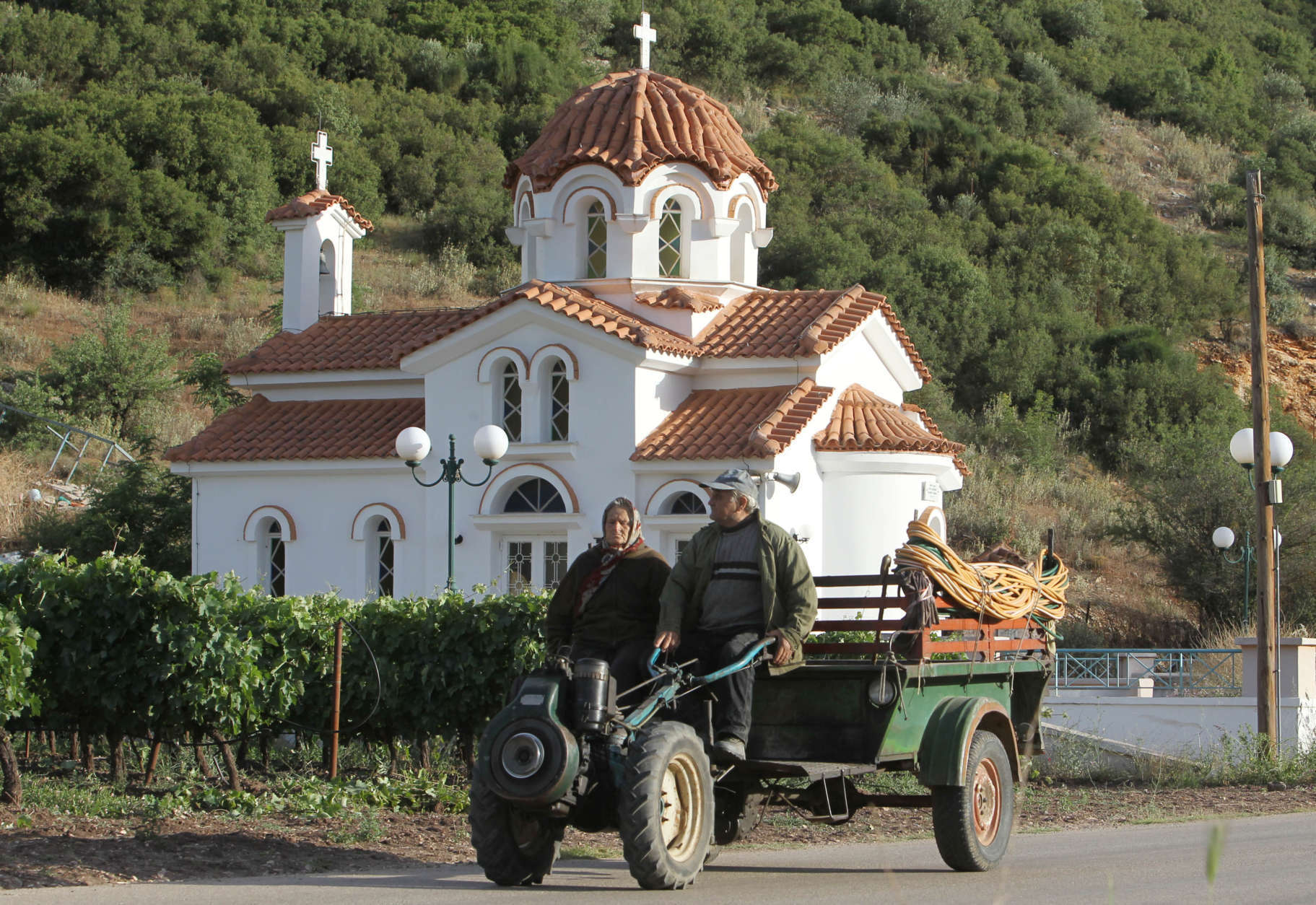 A elderly couple passes a Greek Orthodox with its agricultural machinery next to a vineyard in the town of Nemea about 113 kilometers (70 miles) west of Athens, on Saturday, June 1, 2013. Greece, which is still struggling to stabilize its national debt, is one of the oldest wine-producing regions in the world with the area of Nemea known for the famous red tradition wines. (AP Photo/Thanassis Stavrakis)