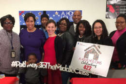 Campaign recipients and D.C. leaders stand in front of Housing Finance Agency goodie bags of supplies for new lease holders at the ‘Home For The Holidays’ lease signing ceremony. (WTOP/Kristi King)