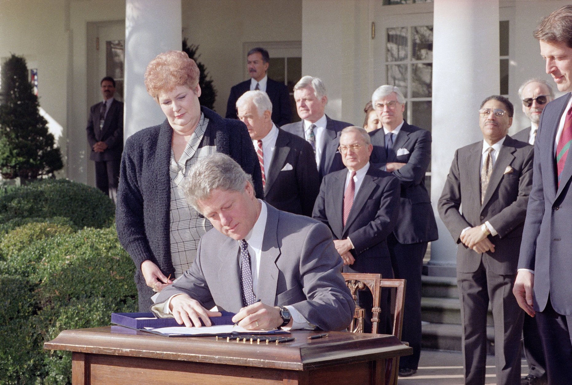 Pres. Clinton signs the Family Leave Bill as Vicki Yandle of Marietta, Ga., looks on in the Rose Garden of the White House, Feb. 5, 1993.  Mrs. Yandle lost her job when she took time off when her daughter was sick.  Behind the president, from left, are: House Speaker Thomas Foley, Sen. Edward M. Kennedy (D-Mass.), Rep. William Ford (D-Mich.), and Sen. Christopher Dodd (D-Conn.) At far right is Vice President Al Gore. (AP Photo/Greg Gibson)