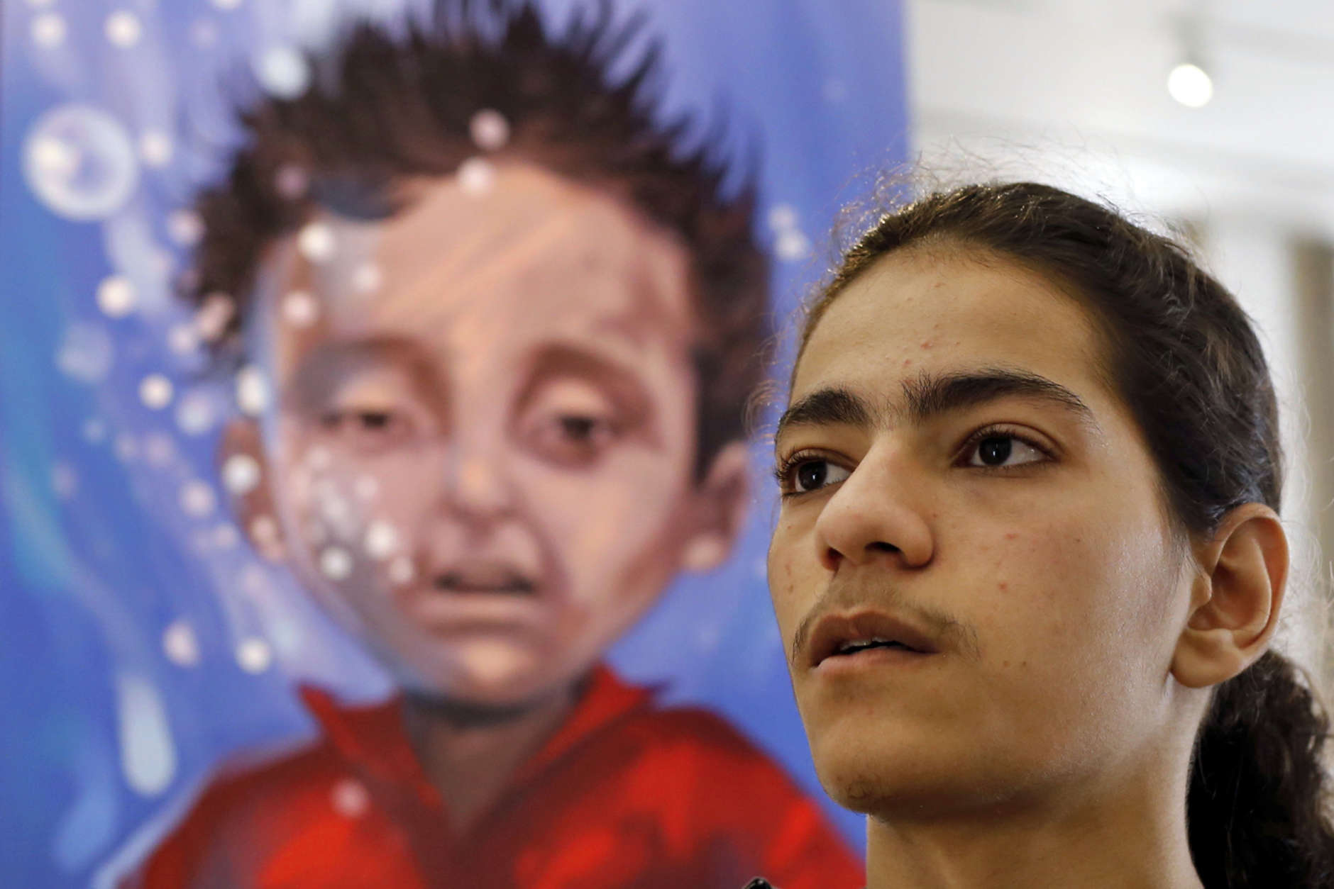 In this Monday, March 12, 2018 photo, Bassel Mokdad, 17, who is paralyzed from a spinal injury incurred during the war in Syria, speaks to The Associated Press, next to a painting of 3-year-old Syrian Aylan Kurdi, who was found dead on a Turkish beach, at the Beit Beirut museum, Beirut, Lebanon. After seven years of war in Syria, the United Nations has one thing to say: Stop the war on children. Of Syria’s estimated 10 million children, 8.6 million are now in dire need of assistance, nearly 6 million children are displaced or living as refugees and about 2.5 million are out of school. (AP Photo/Bilal Hussein)