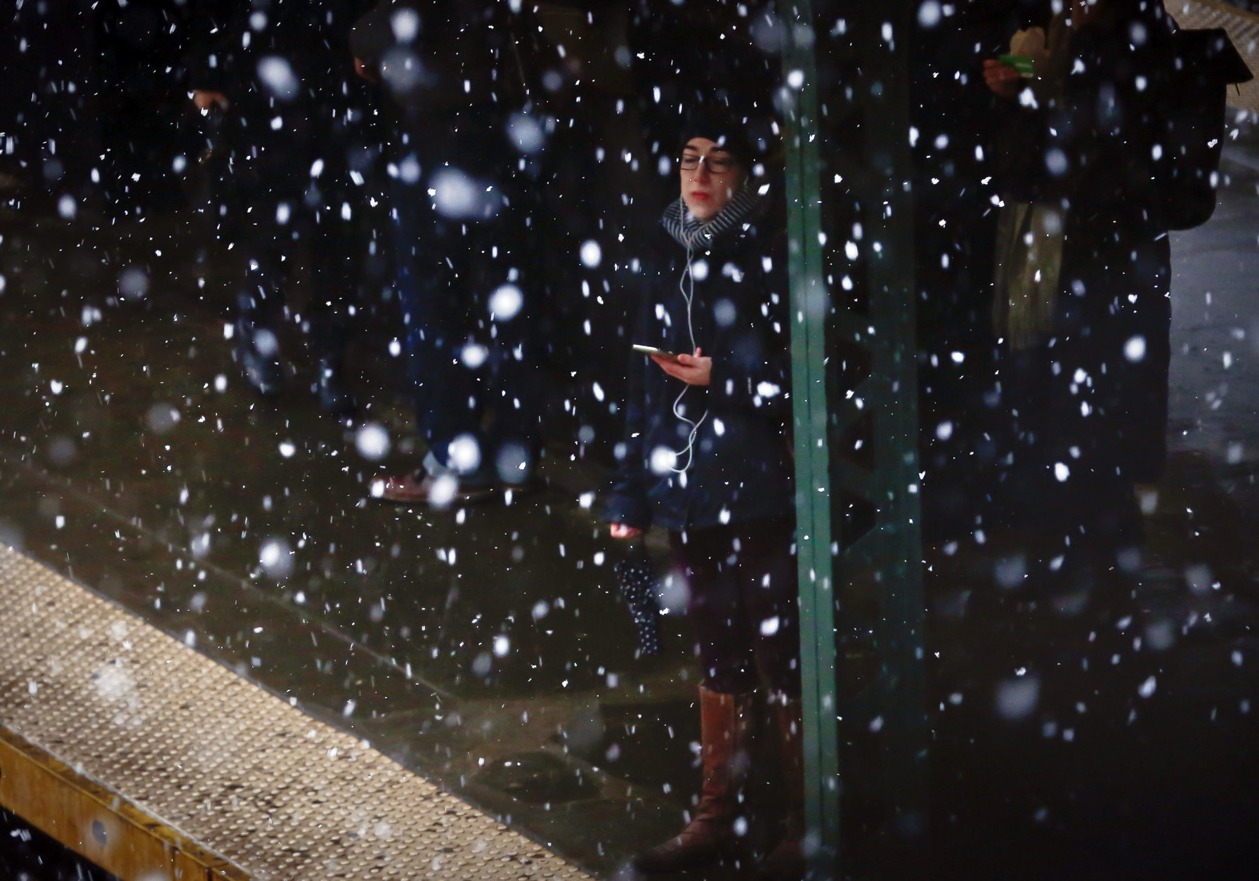 A commuter, with ear plugs tethered to a phone, is sheltered from snowfall while waiting on a subway platform, Tuesday March 13, 2018, in the Brooklyn borough of New York. The National Weather Service predicted between two and five inches of snow in the city and surrounding areas, where a winter weather advisory was in effect. (AP Photo/Bebeto Matthews)