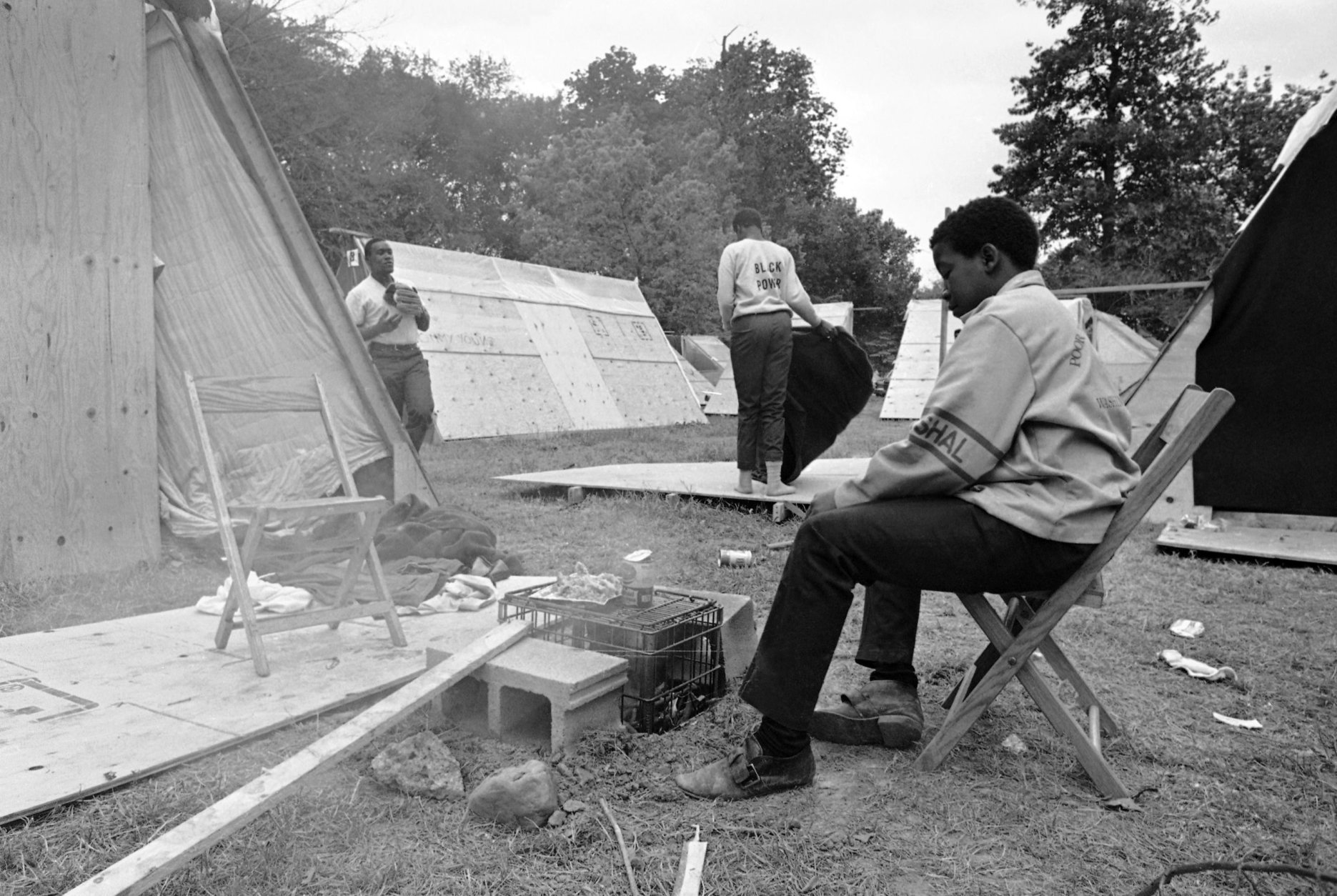 Monroe Johnson, 13, of Milwaukee, Wisconsin, cooks his mid-day meal outdoors in "Resurrection City," the camp site of the Poor People's Campaign in Washington, May 20, 1968. Another resident shakes a blanket amid the plywood shelters which make up the camp. (AP Photo/Bob Schutz)