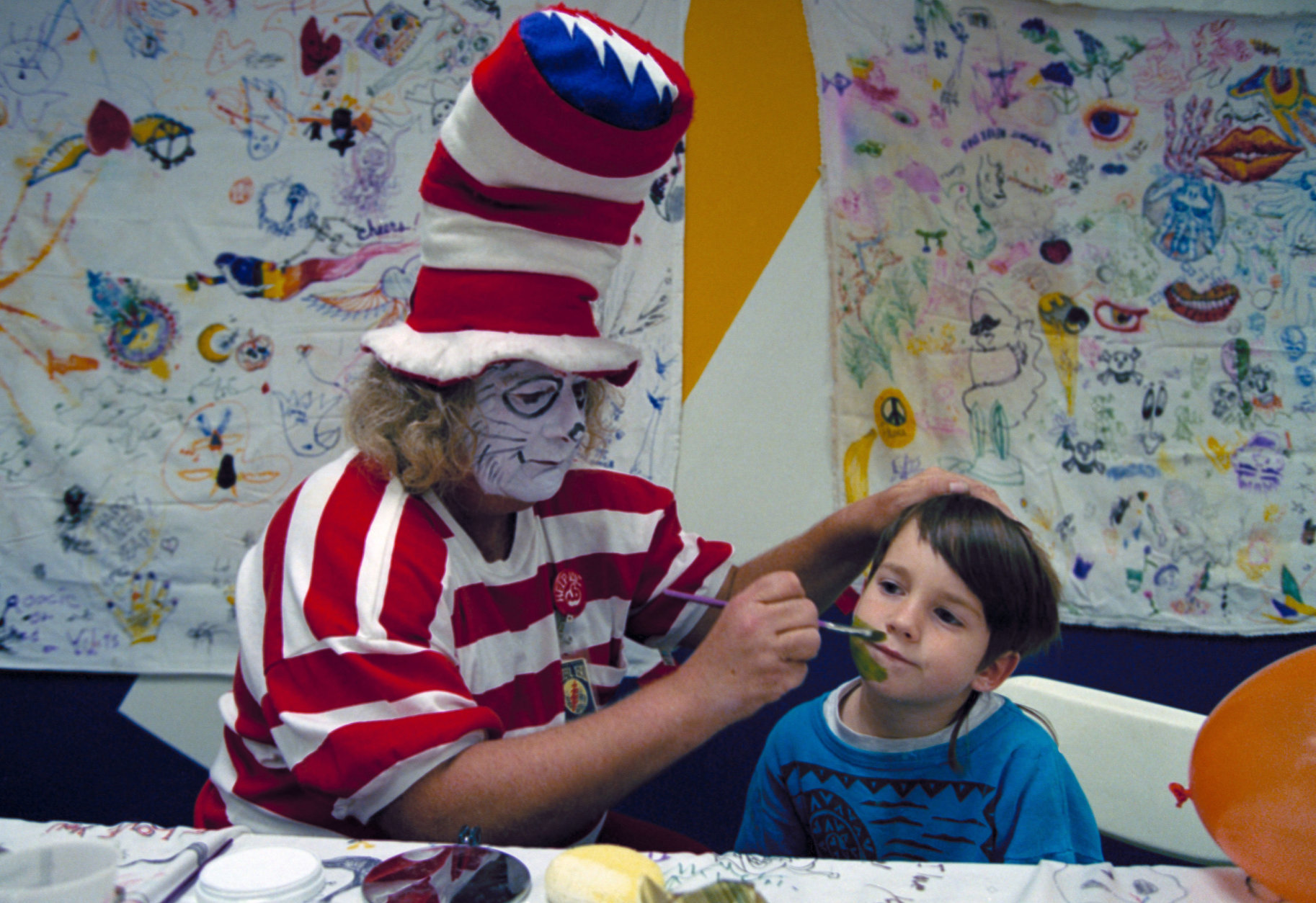 Wavy Gravy, costumed as The Cat in the Hat, paints the face of 5-year-Old Jesse James Nugent backstage at the Oakland Coliseum during a Grateful Dead concer February 14, 1993.  Gravy, a backstage presence since the 1960s, entertains kids in the daycare room set up for the children of the band members and crew. (AP Photo/Eric Risberg)