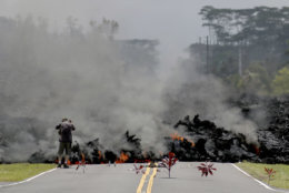 Lava burns across a road in the Leilani Estates subdivision as an unidentified person takes pictures of the flow, Saturday, May 5, 2018 near Pahoa, Hawaii. Offerings of Hawaiian ti leaves, rocks and cans to the fire goddess Pele lie in the street in front of the lava. (AP Photo/Caleb Jones)