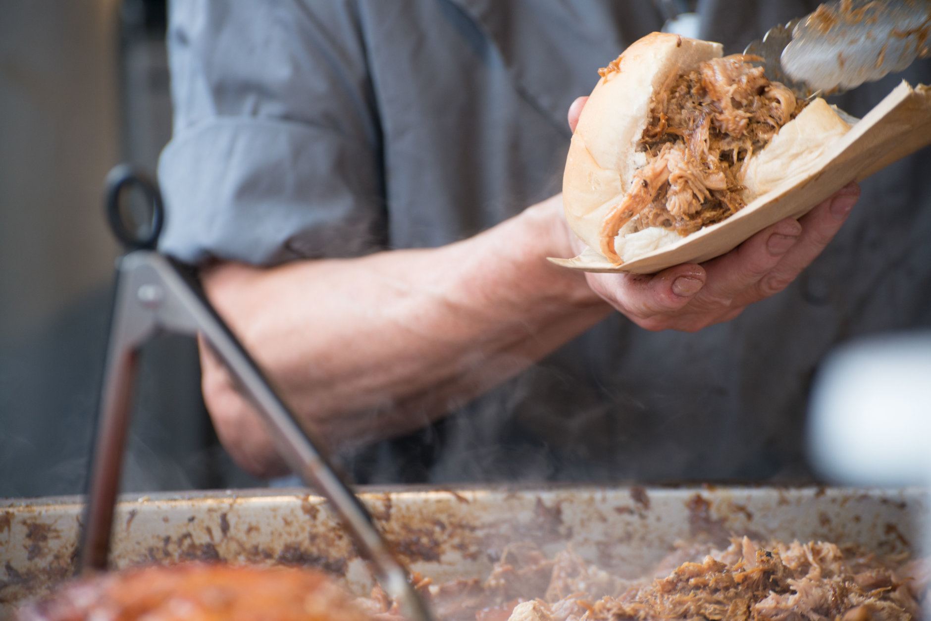 Man in grey T shirt serving pulled pork street food holding sandwich with rind and serving tongs in the foreground