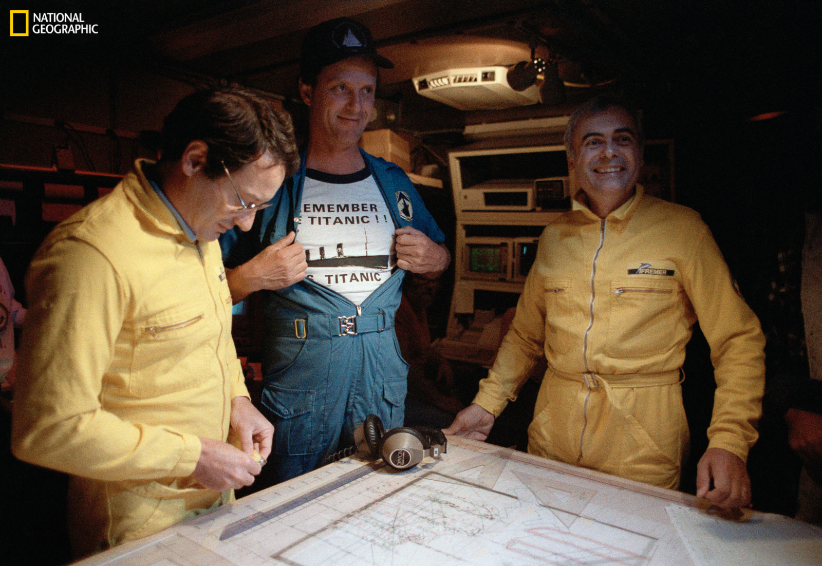 Titanic expedition members pore over blueprints of the R.M.S. Titanic.  A joint expedition by the Woods Hole Oceanographic Institution and the Institut Franca is de Recherches pour L'exploitation des Mers was formed to find the Titanic's final resting place.  The chief scientist of the Woods Hole contingent was Robert D. Ballard, here proudly displaying a commemorative expedition t-shirt. (Emory Kristof/National Geographic)
