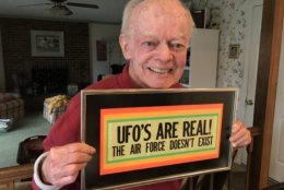 Shea holds a funny sign that used to hang in his office. It was given to him by journalist and UFO skeptic Phil Klass. (WTOP/Michelle Basch)