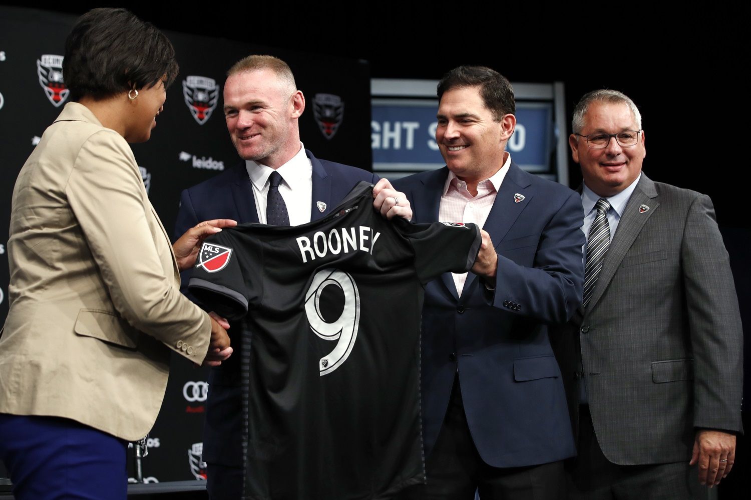 District of Columbia Mayor Muriel Bowser, left, shakes hands with English soccer star Wayne Rooney, the all-time leading scorer for England's national team and Manchester United in the Premier League, as he holds up his new jersey next to MLS team D.C. United Managing Partner and CEO Jason Levien, and Dave Kasper, General Manager and Vice President of Soccer Operations, right, during a news conference announcing Rooney's signing with D.C. United, Monday, July 2, 2018, at the Newseum in Washington. (AP Photo/Jacquelyn Martin)