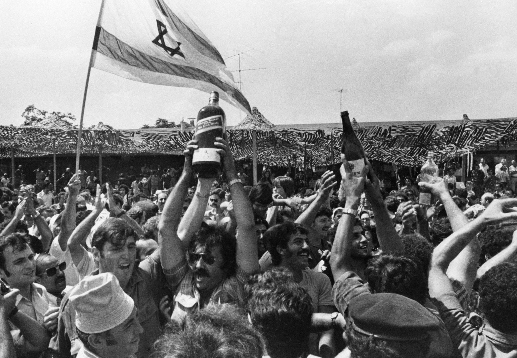 Some of the hundreds of relatives and well-wishers who came to Ben-Gurion Airport near Tel Aviv, July 4, 1976 for return of hijacked Air France passengers rescued in night raid Uganda, where they were held for a week raise Israeli flag and bottles of drink in toast. (AP Photo/Shmuel Rachmani)