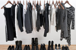 Women's clothing hanging from a metal bar and shoes on the hardwood floor beneath them.  The clothes and shoes are primarily black with some white and gray.  A white wall is in the background.