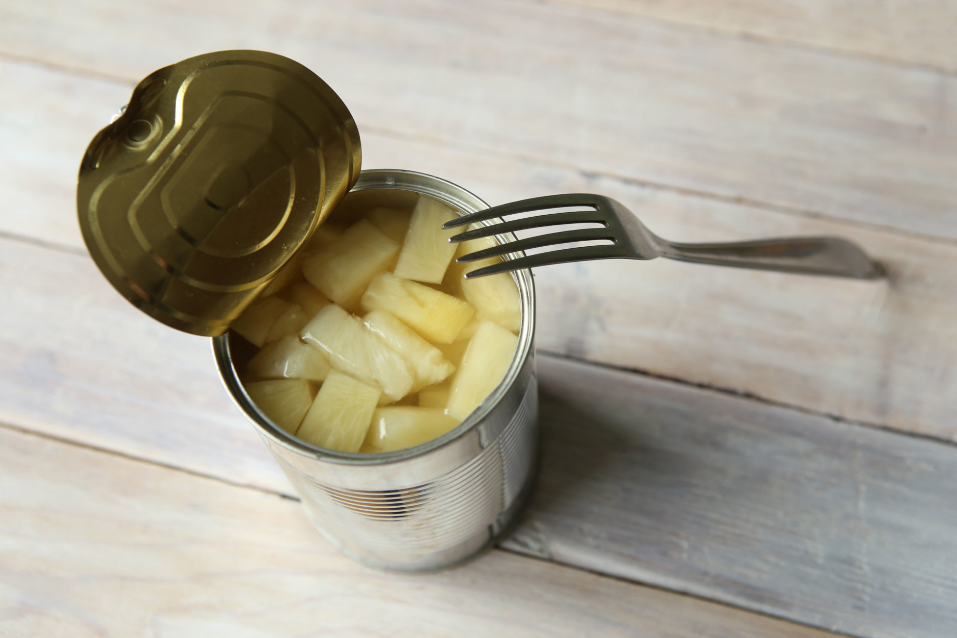 Canned pineapple in can with fork on wooden table.