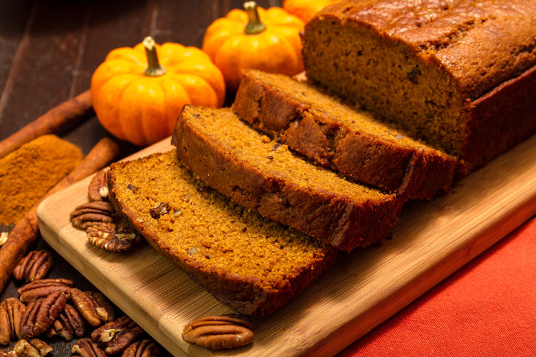 Sliced pumpkin bread loaf sitting on wooden cutting board with pecan nuts and cinnamon spices