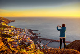 Young female traveler in blue jacket photographing Santa Cruz city standing on the top of the mountain on La Palma island in the morning