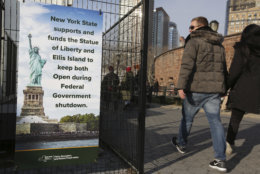 Tourists walk past a sign near the embarkation point for the Statue of Liberty in New York, Thursday, Dec. 27, 2018. The Statue of Liberty and Ellis Island will remain open despite the ongoing partial government shutdown, even as some national parks and monuments close down, according to New York Gov. Andrew Cuomo. (AP Photo/Seth Wenig)