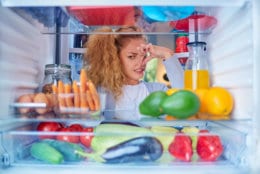 Woman standing in front of opened fridge and holding up to her nose because of bad smell. Picture taken from the inside of fridge full of groceries.